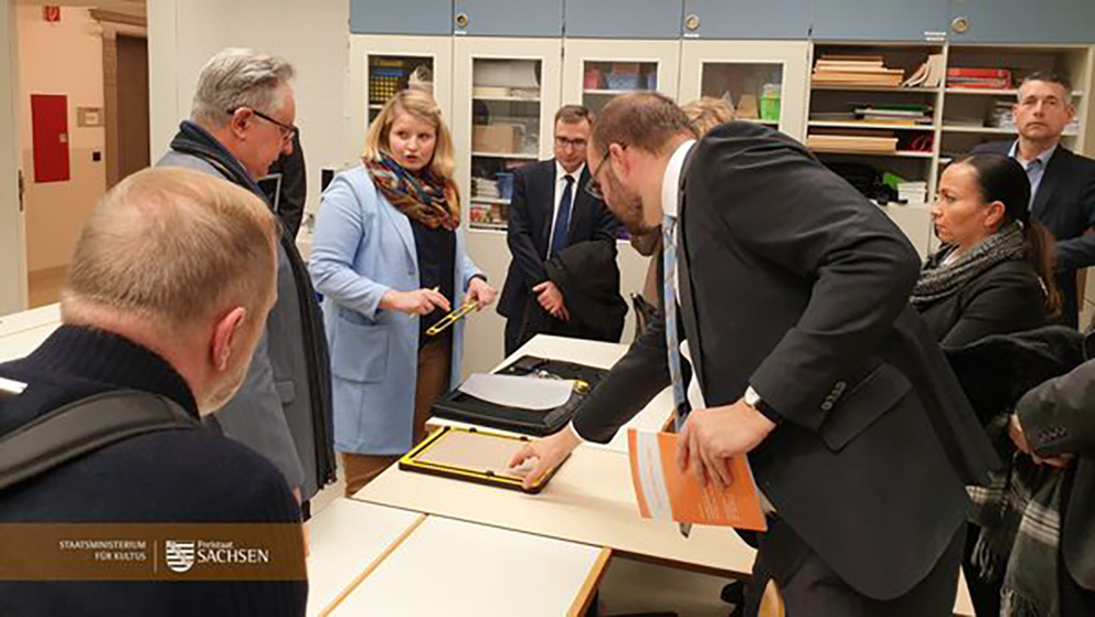 Bildbeschreibung: Die Leiterin des Kompetenz- und Medienzentrums Frau Böttcher erklärt der polnischen Delegation und den sächsischen Vertretern das Zeichengerät Tactipad. Alle Personen stehen um einen Schülertisch auf dem das Zeichnematerial liegt. Staatsminister Piwarz fühlt gerade mit einer Hand die Unebenheiten einer gezeichneten Linie auf der Folie.