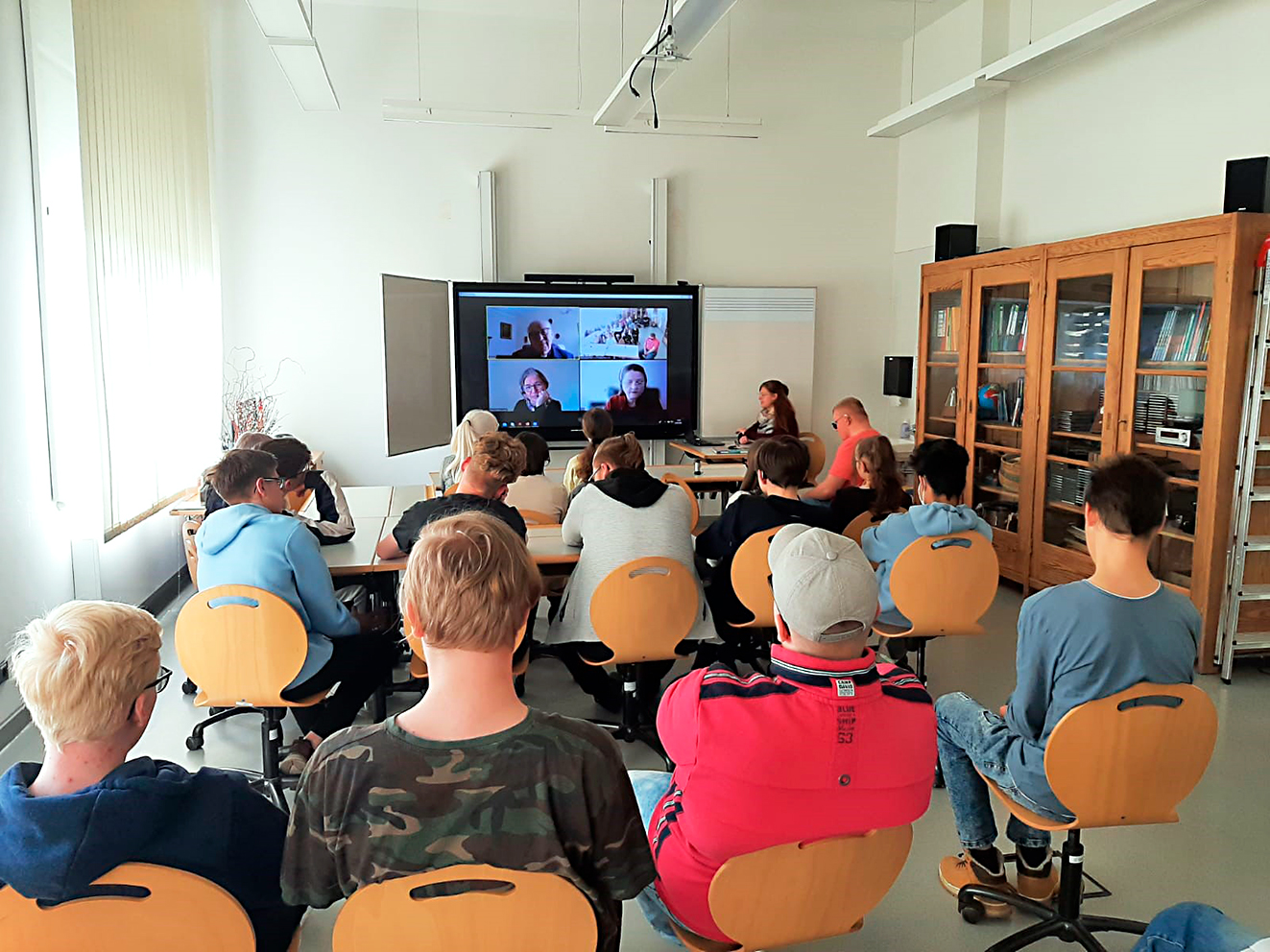 Das Bild zeigt Schülerinnen und Schüler von hinten. Sie sitzen auf Stühlen in einem Klassenzimmer und schauen Richtung digitaler Tafel. Auf dieser sieht man die Bilder von drei einzelnen Personen sowie ein Bild der Schülerinnen und Schüler in einer Videokonferenz.